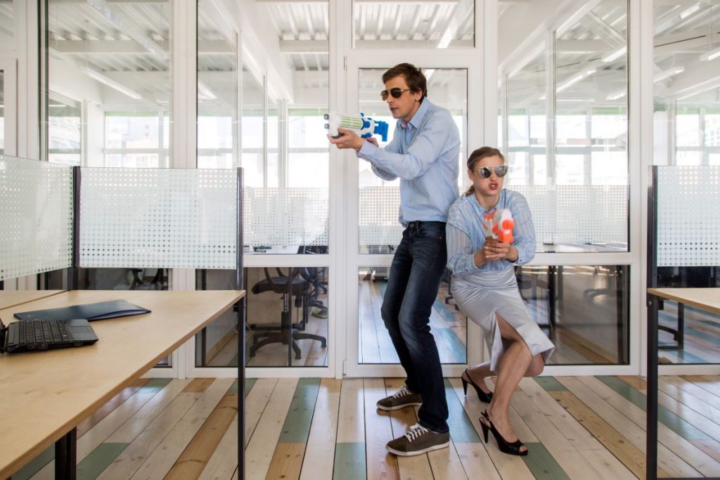 Mann und Frau in formeller Kleidung stehen Rücken an Rücken und zielen auf Nerf Gun Spielzeugpistolen, während sie sich im Büro amüsieren