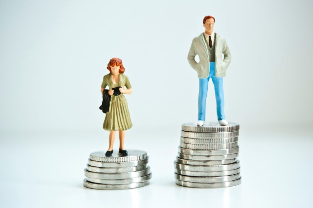 Das Bild stellt einen Mann dar, der auf einem höheren Stapel Geld steht als eine Frau, was symbolisch den Lohnunterschied zwischen den Geschlechtern darstellt.